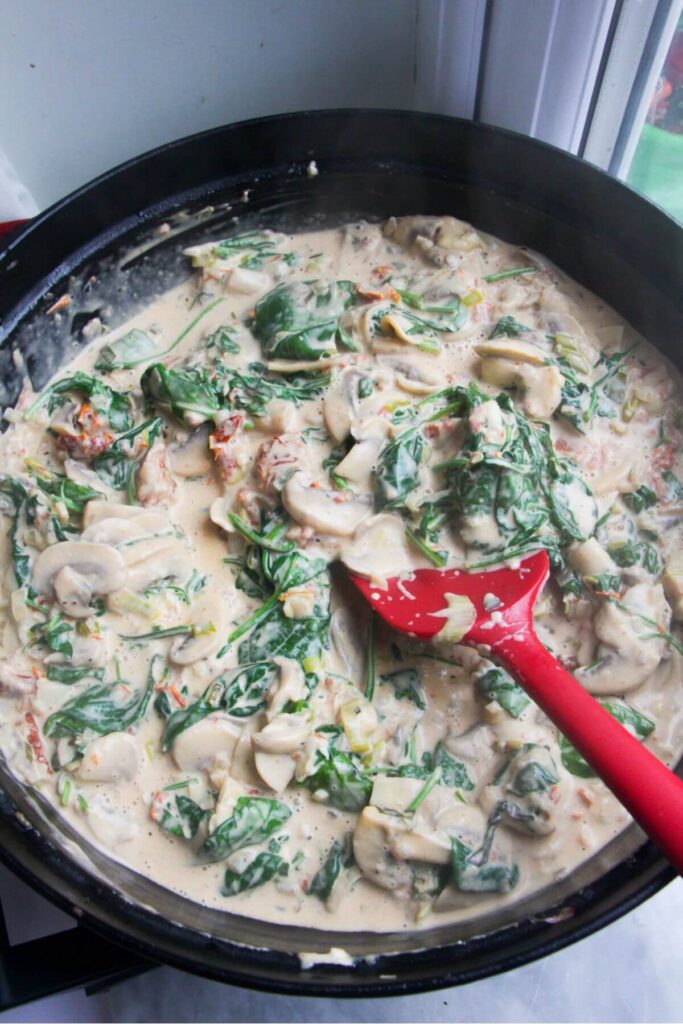 Red spatula stirring spinach leaves into creamy mushroom sauce in a large pan.