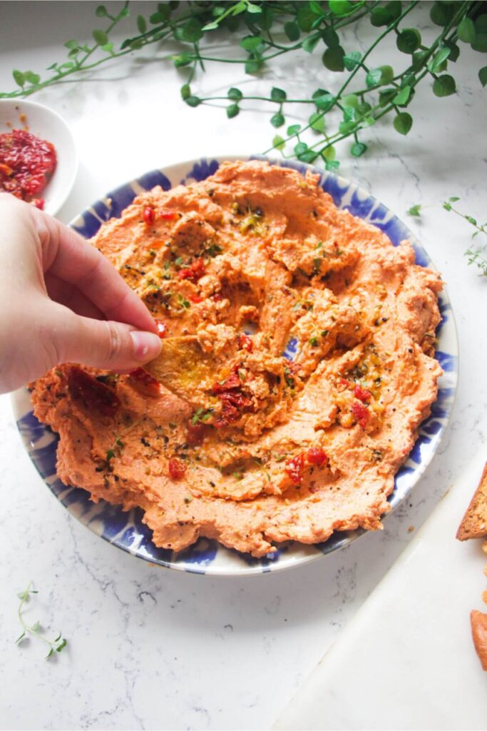Hand dipping pita chip in sundried tomato feta dip on a small blue and white plate.