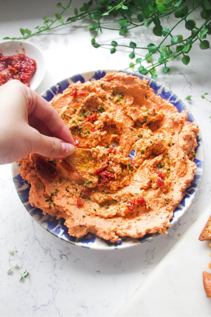 Hand dipping pita chip in sundried tomato feta dip on a small blue and white plate.