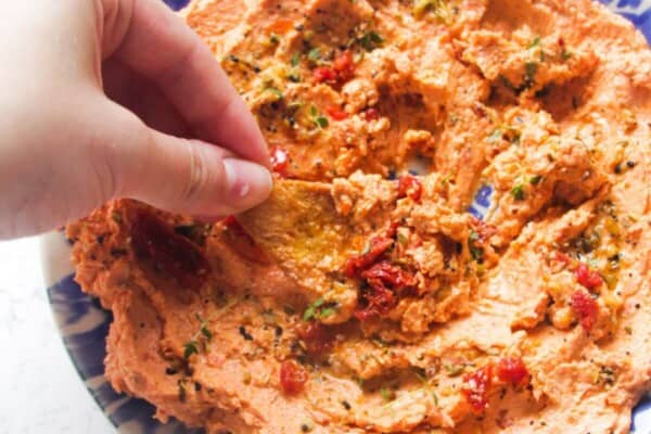 Hand dipping pita chip in sundried tomato feta dip on a small blue and white plate.