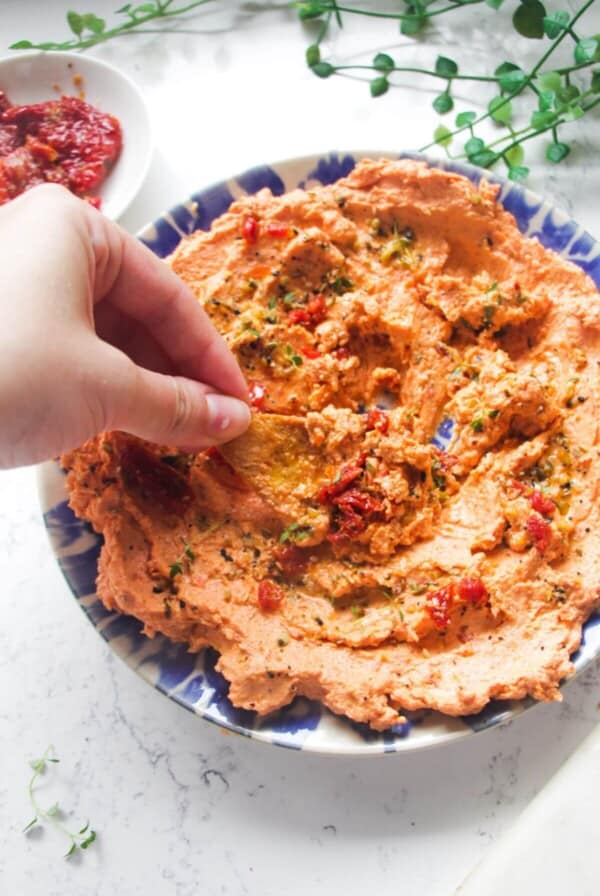 Hand dipping pita chip in sundried tomato feta dip on a small blue and white plate.
