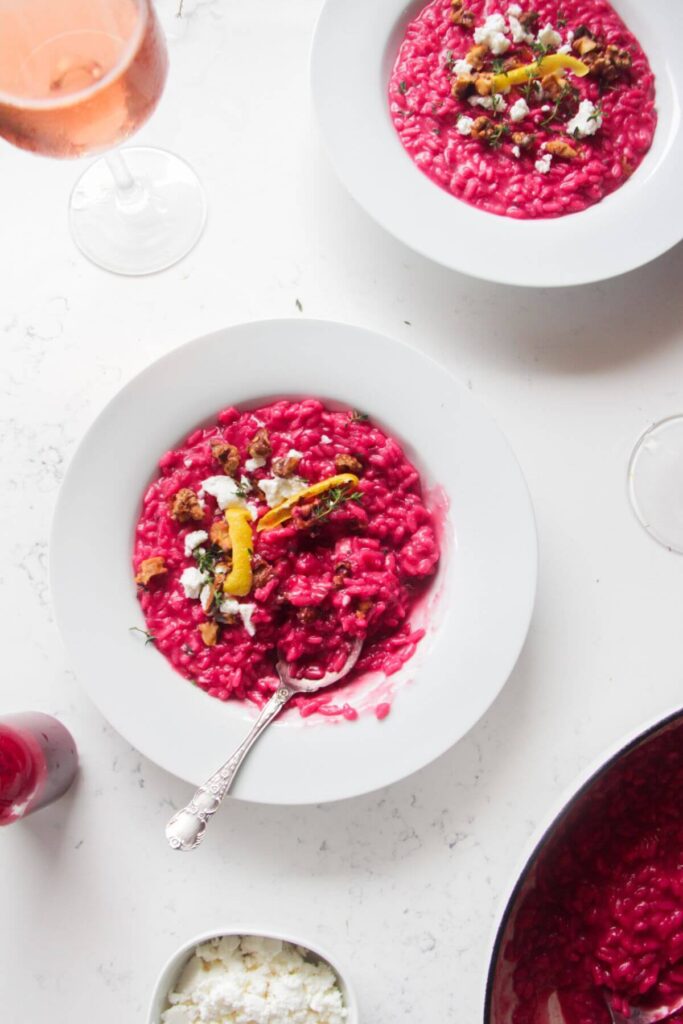 Bright pink beetroot risotto in a white bowl, with feta, lemon peel and walnuts on top with a silver spoon.