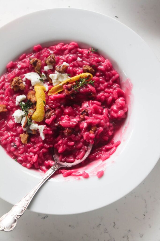 Bright pink beetroot risotto in a white bowl, with feta, lemon peel and walnuts on top with a silver spoon.