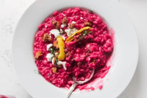 Bright pink beetroot risotto in a white bowl, with feta, lemon peel and walnuts on top with a silver spoon.