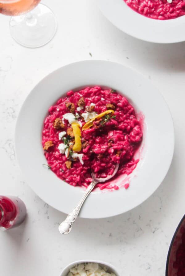 Bright pink beetroot risotto in a white bowl, with feta, lemon peel and walnuts on top with a silver spoon.