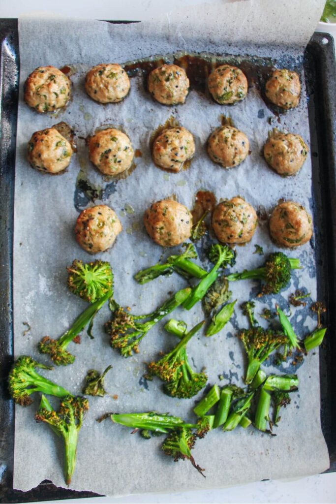 Cooked chicken meatballs and charred broccolini on a lined oven tray.