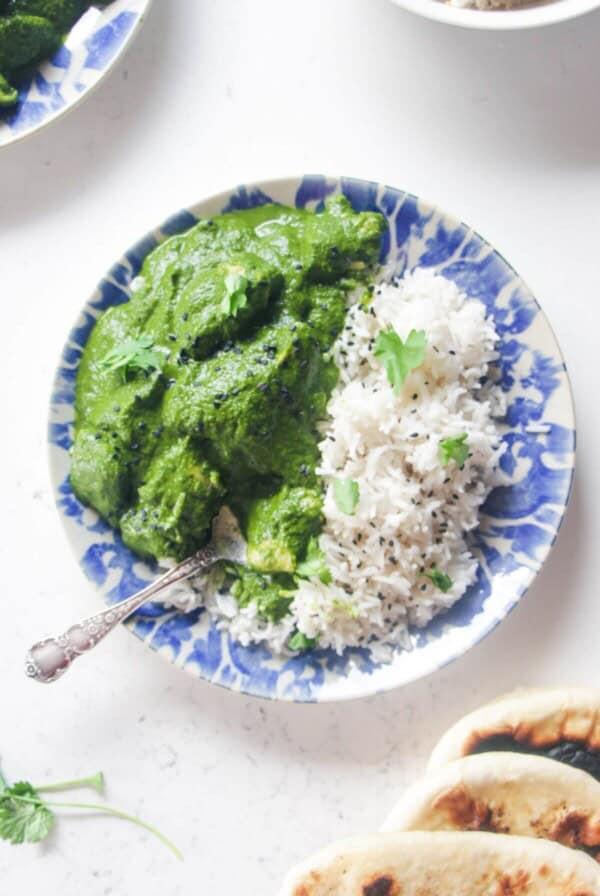 Chicken saag on a white and blue plate with rice, with more curry and naan on the side.