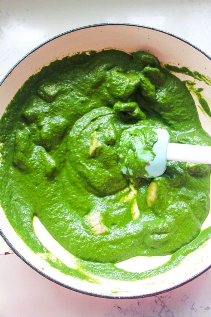 Diced chicken being stirred through bright green saag sauce in a white skillet.
