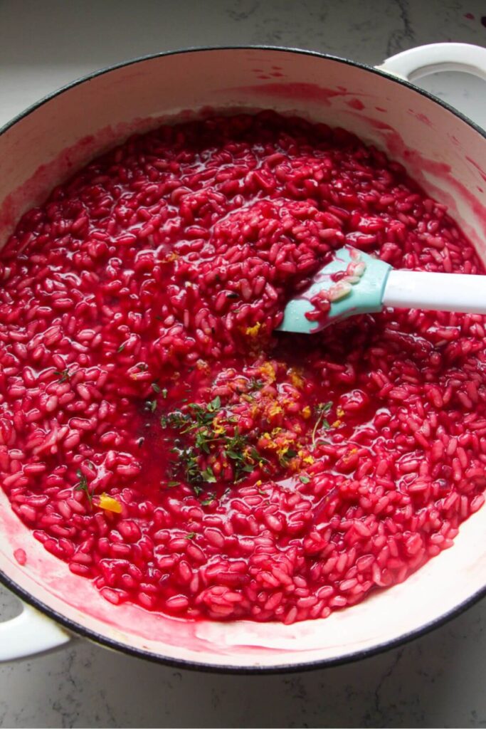 Blue and white spatula stirring thyme and lemon zest through bright pink beetroot risotto.
