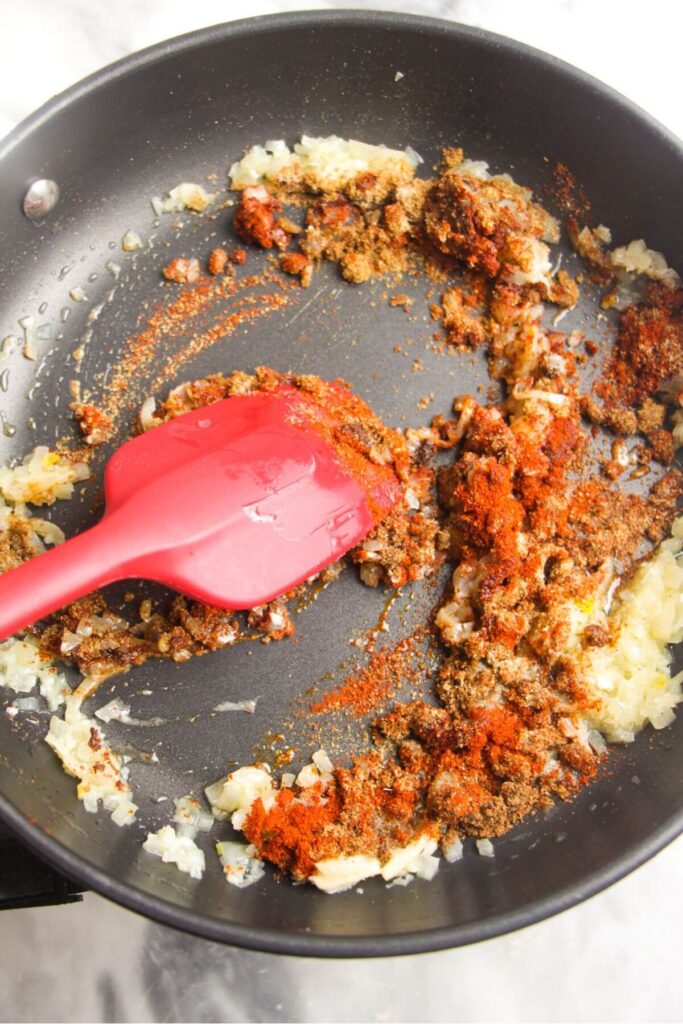 Spices and diced onion stirred by a red spatula in a frying pan.