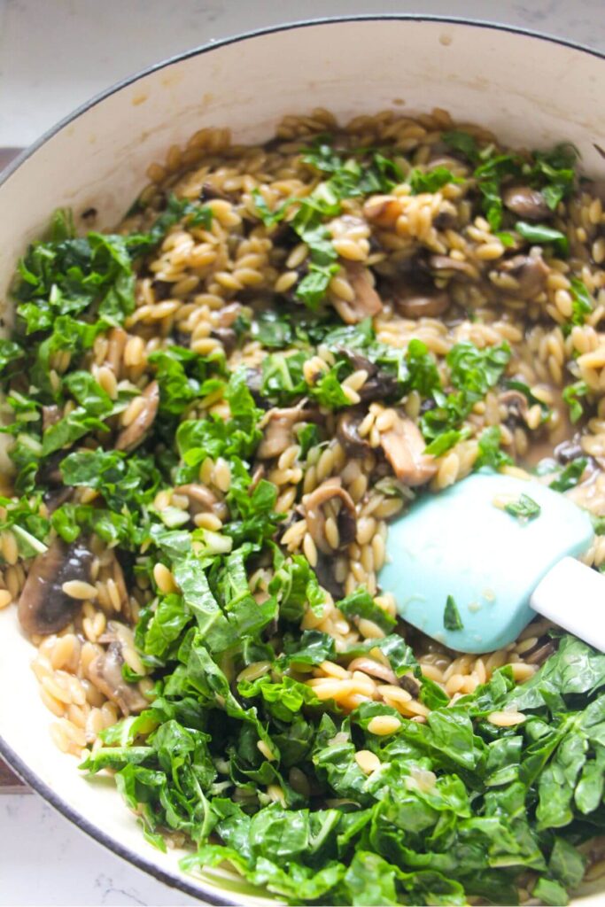Chopped spinach being stirred through mushroom orzo in a large white skillet.