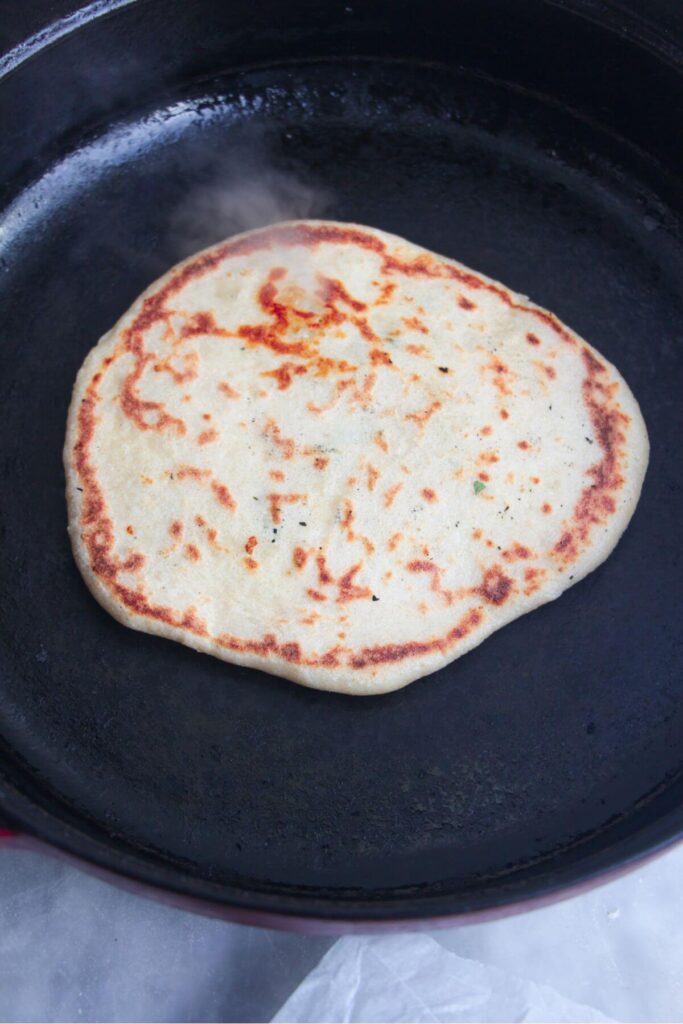 Golden speckled cheese naan cooking in a large black skillet.