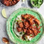 Chicken rogan josh on a bed of white rice on a green plate, with more curry in a small silver dish on the side.