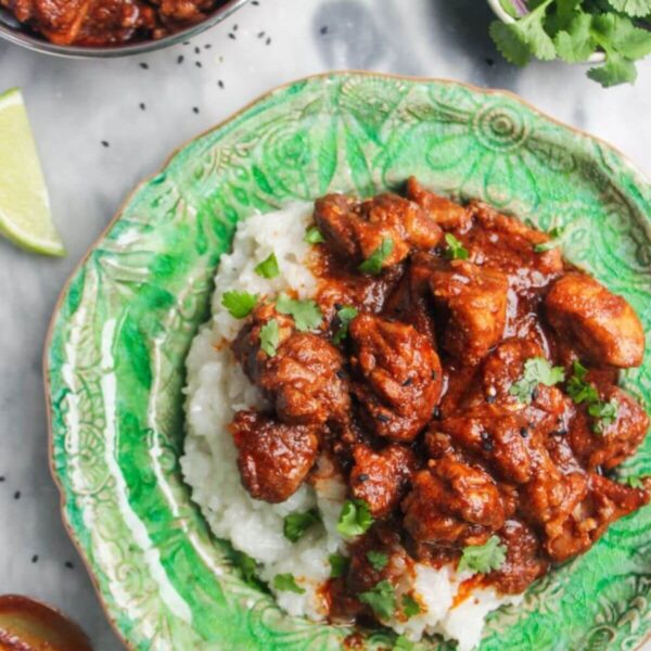 Chicken rogan josh on a bed of white rice on a green plate, with more curry in a small silver dish on the side.