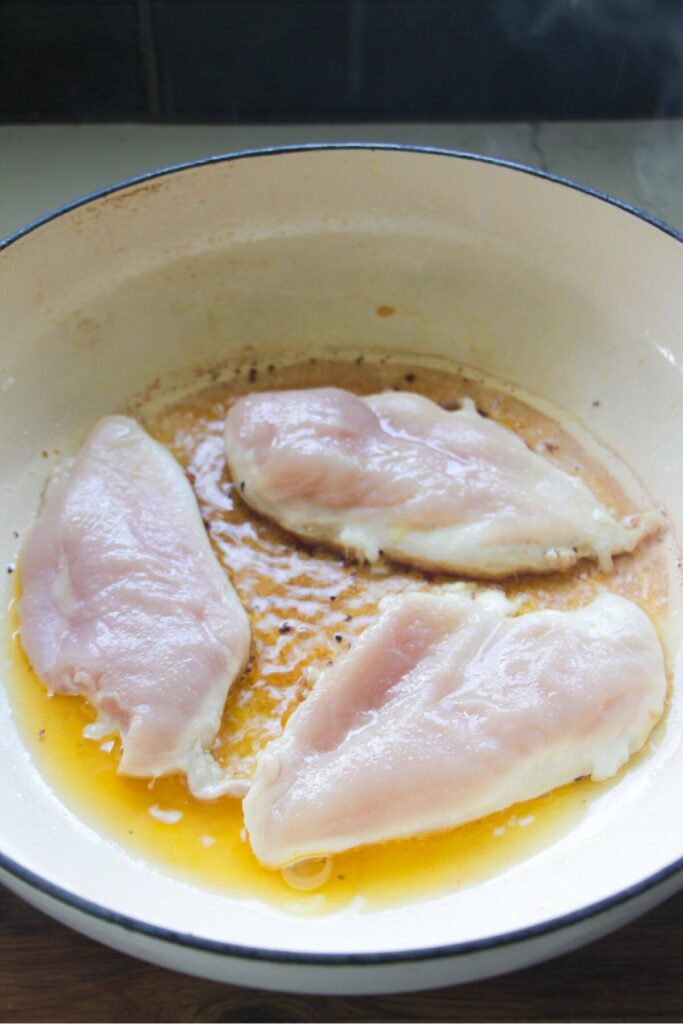 Three chicken breasts cooking in a large white skillet.