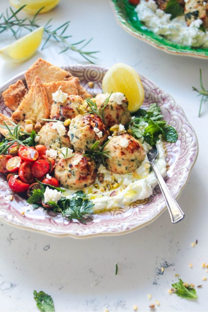 Chicken meatballs, tomatoes, on tzatziki with pita chips on a pink plate.