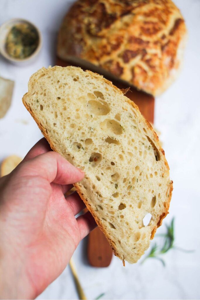 Hand holding up a slice of rosemary parmesan bread with the rest of the loaf in the background.