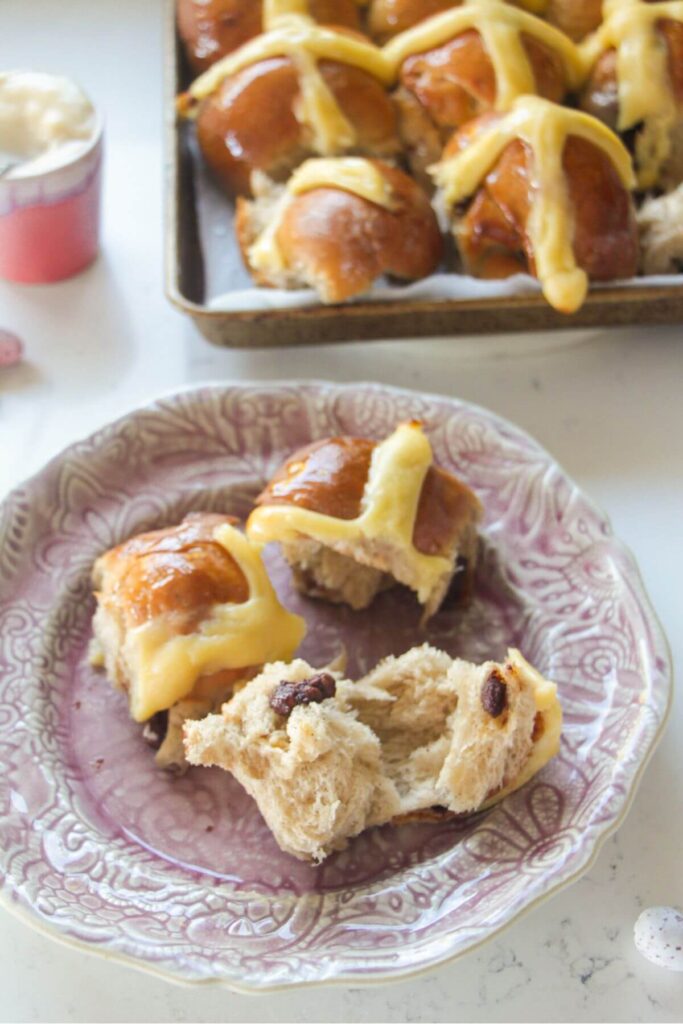 Torn open mini egg hot cross bun on a pink plate with more hot cross buns in the background.