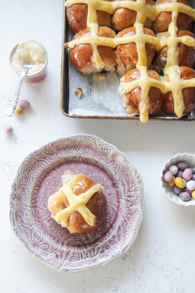 Mini egg hot cross bun on a small pink plate with more hot cross buns in the background.