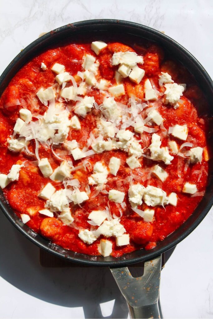 Cheese added on top of gnocchi in tomato sauce in a small frying pan.