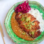 Sliced, breaded chicken breast on top of rice, curry sauce and pickled red cabbage on a green plate.