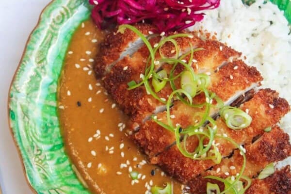 Sliced, breaded chicken breast on top of rice, curry sauce and pickled red cabbage on a green plate.