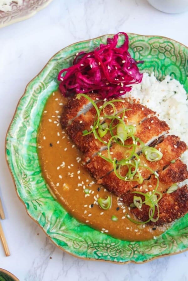 Sliced, breaded chicken breast on top of rice, curry sauce and pickled red cabbage on a green plate.
