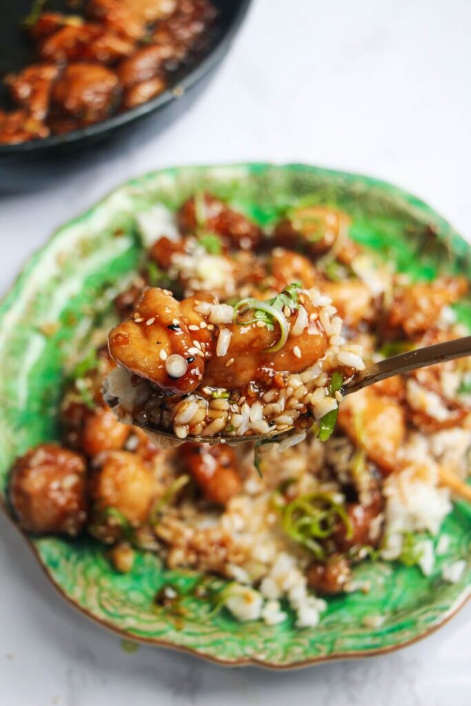 Close up of rice and honey sesame chicken on a spoon with more chicken in the background.