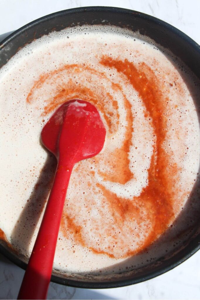 Red spatula stirring coconut milk through tomato sauce in a frying pan.