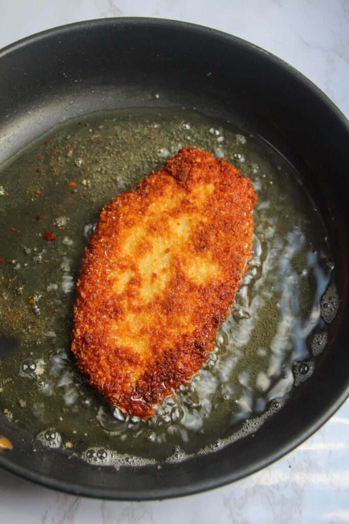Breaded golden chicken breast cooking in oil in a small frying pan.