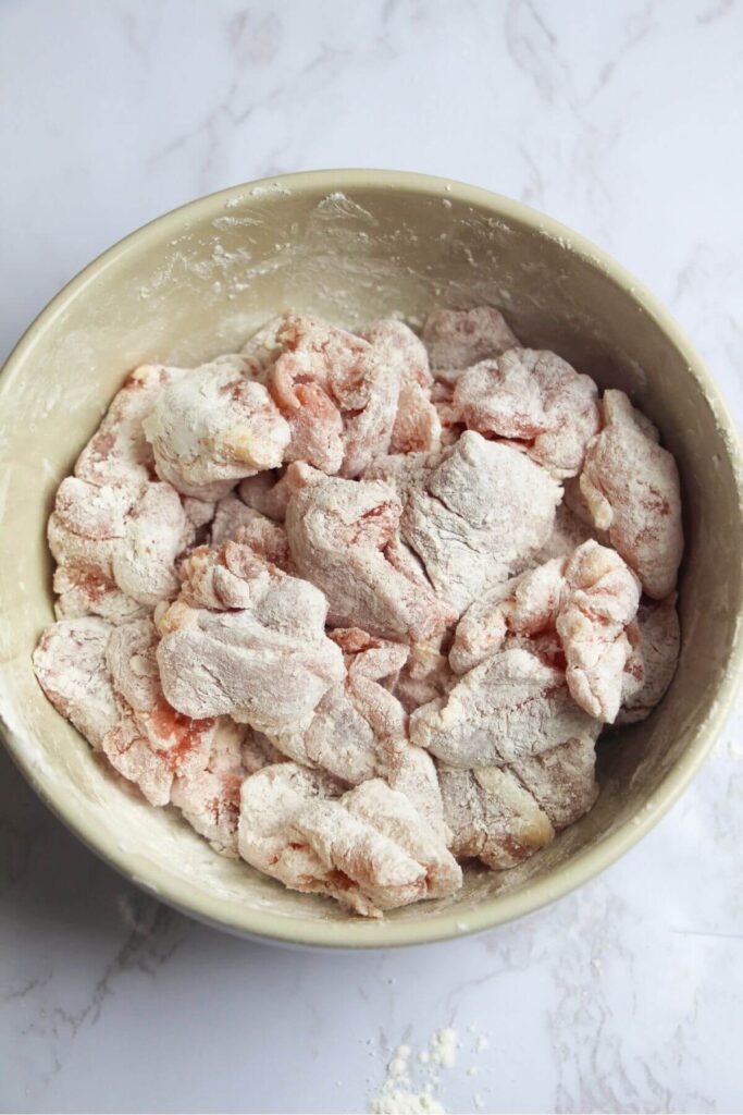 Chicken thigh pieces tossed through cornstarch in a small white bowl.