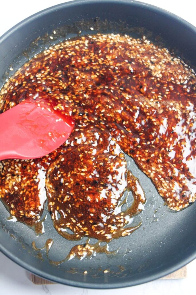 Red spatula stirring honey sesame sauce in a frying pan.