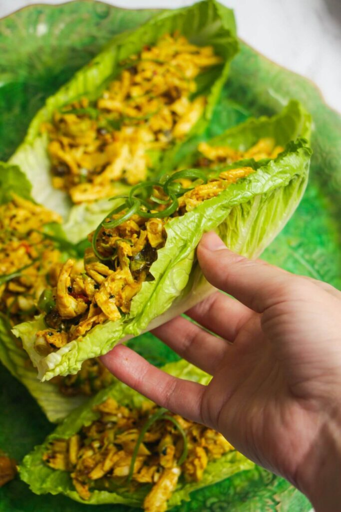 Hand holding a lettuce leaf with coronation chicken inside with more coronation chicken in the background.