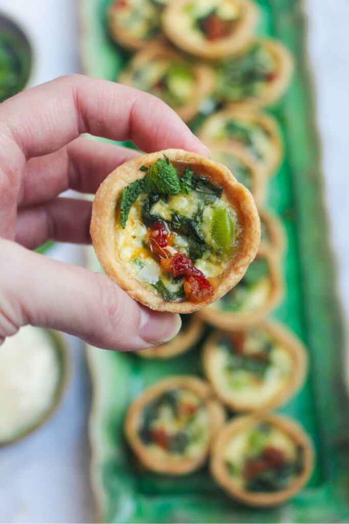 Hand holding up a mini coronation quiche with more mini quiches in the background.