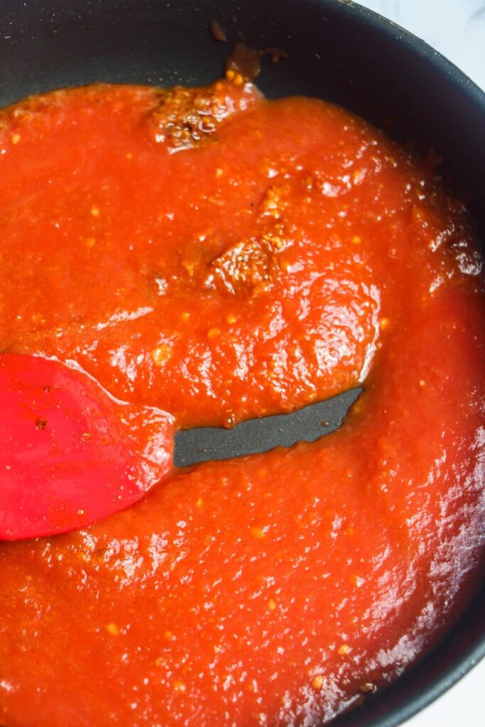 Red spatula stirring tomato sauce in a black pan.