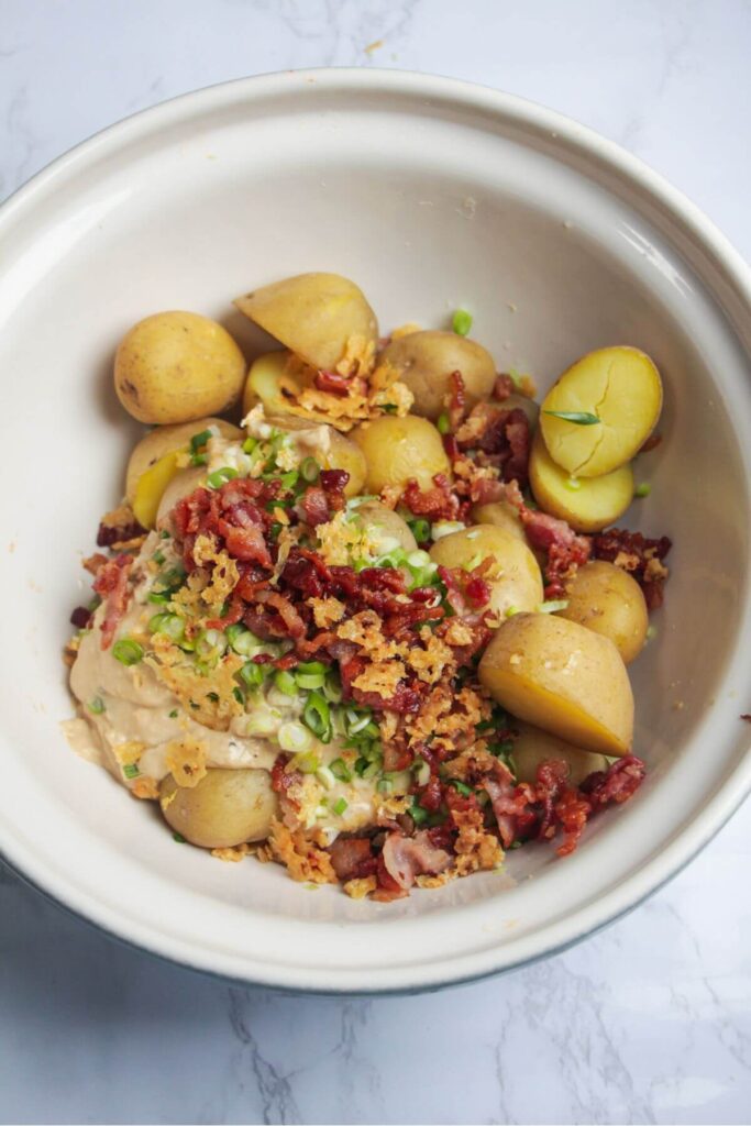 Cooked potatoes, bacon, scallions, crispy parmesan and dressing in a large white bowl.