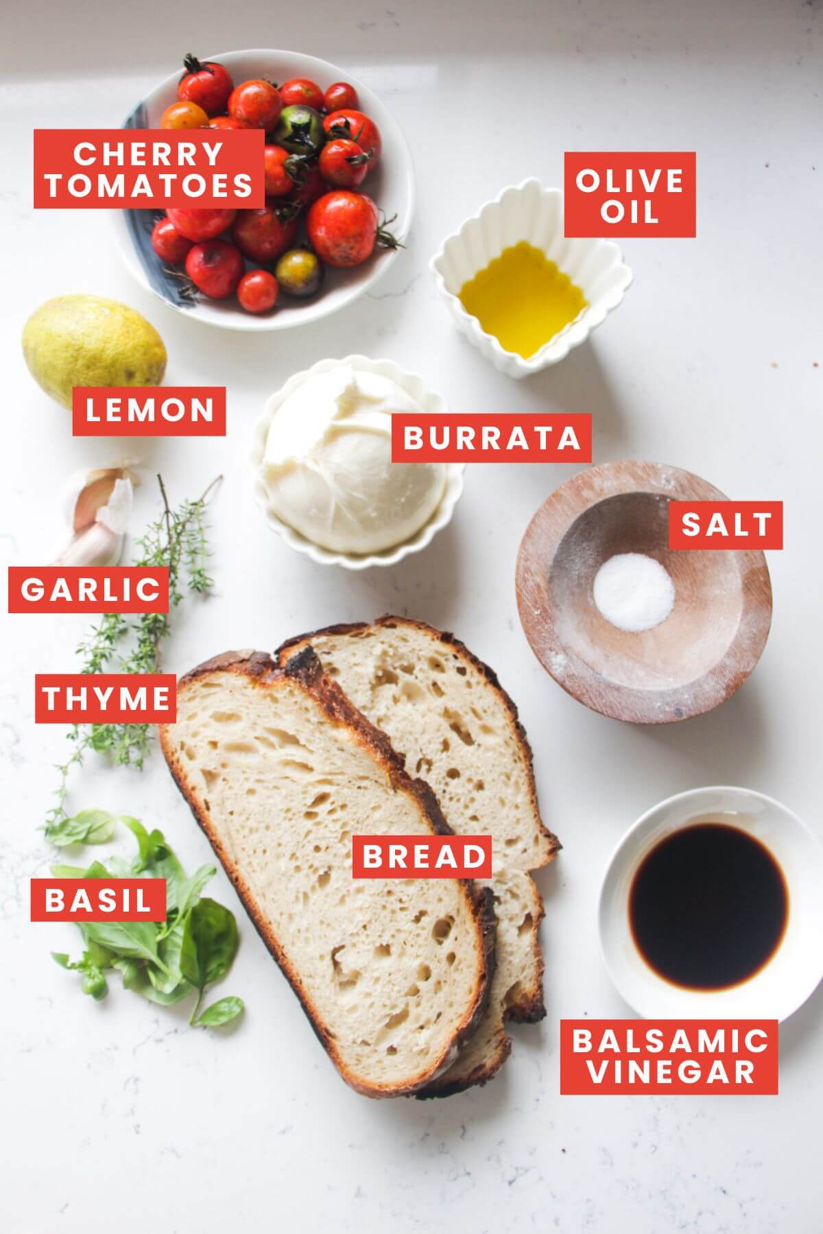 Ingredients for roasted tomato and burrata toast laid out on a white marble background and labelled.