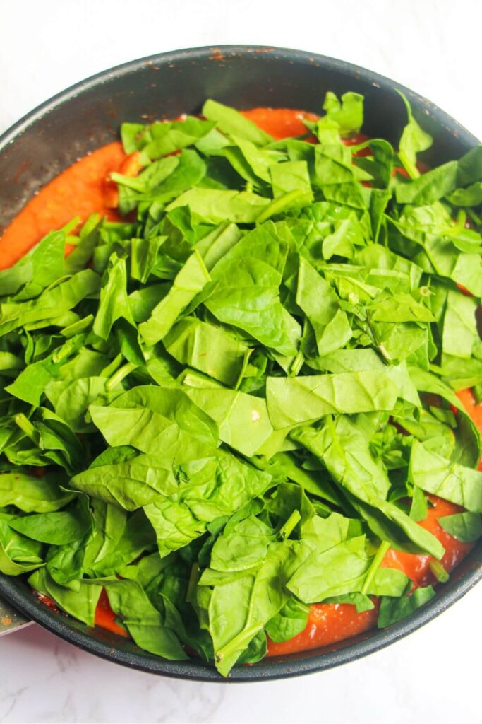 Green spinach leaves added to tomato sauce in a frying pan.