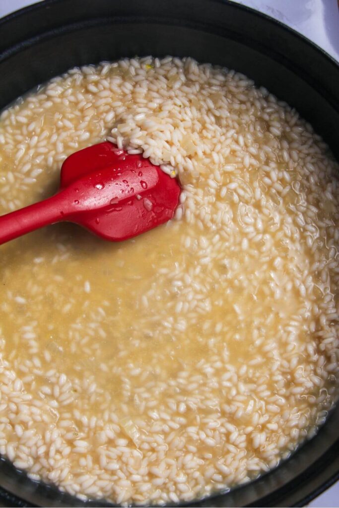 Red spatula stirring stock into rice in a large black skillet.