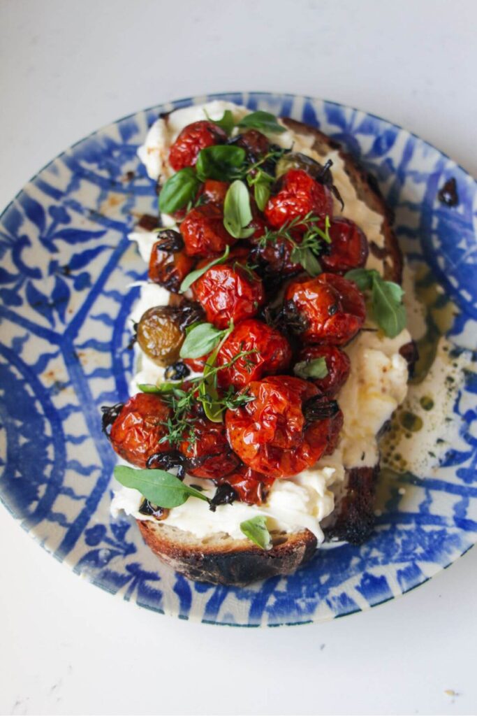 Close up of roasted cherry tomatoes on top of burrata on toast, with green herbs.