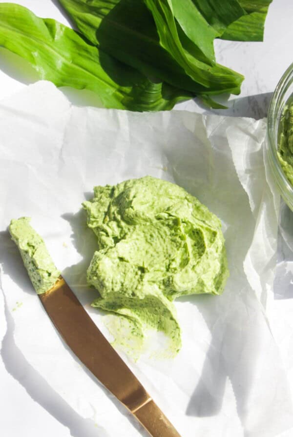 Wild garlic butter on a piece of baking paper with more wild garlic butter in a small glass bowl, with a gold knife on the side.