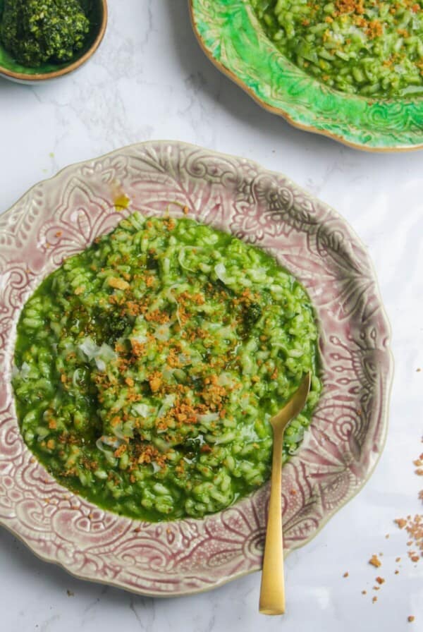 Wild garlic risotto topped with crispy breadcrumbs on a small pink plate, with another plate of risotto in the background.