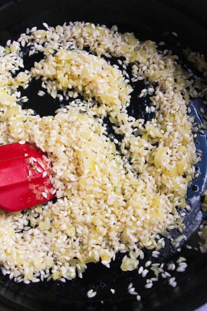 Red spatula stirring rice through onion and garlic in a large black skillet.