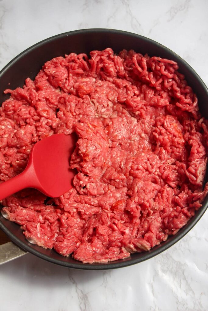 Ground beef added to a frying pan and being stirred with a red spatula.