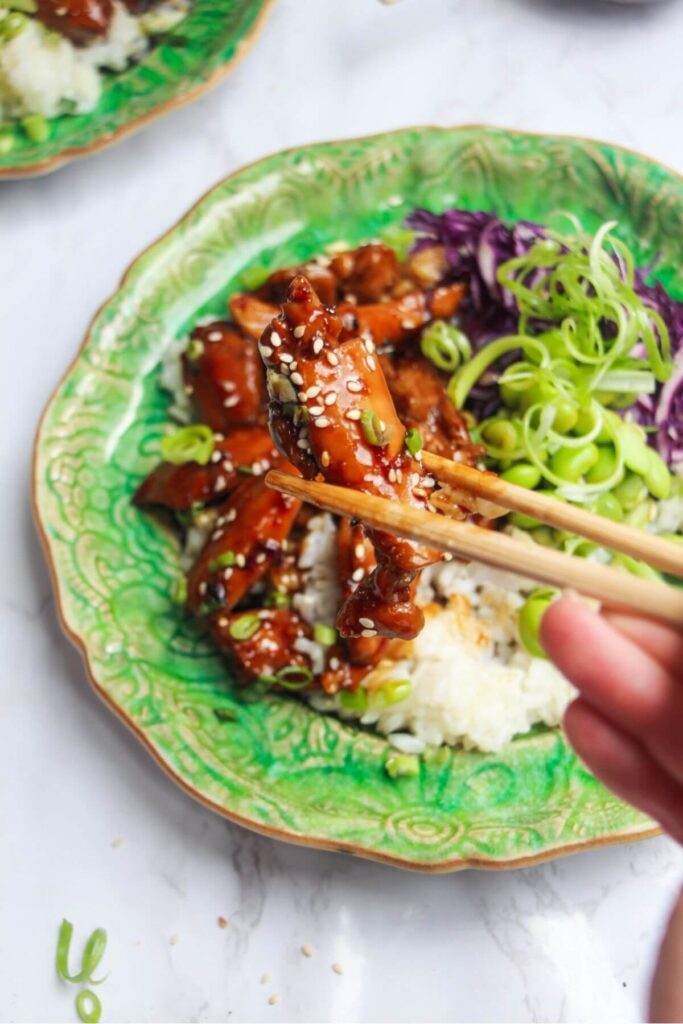 Hands holding chopsticks with a piece of teriyaki chicken in them, with teriyaki chicken bowl in the background.