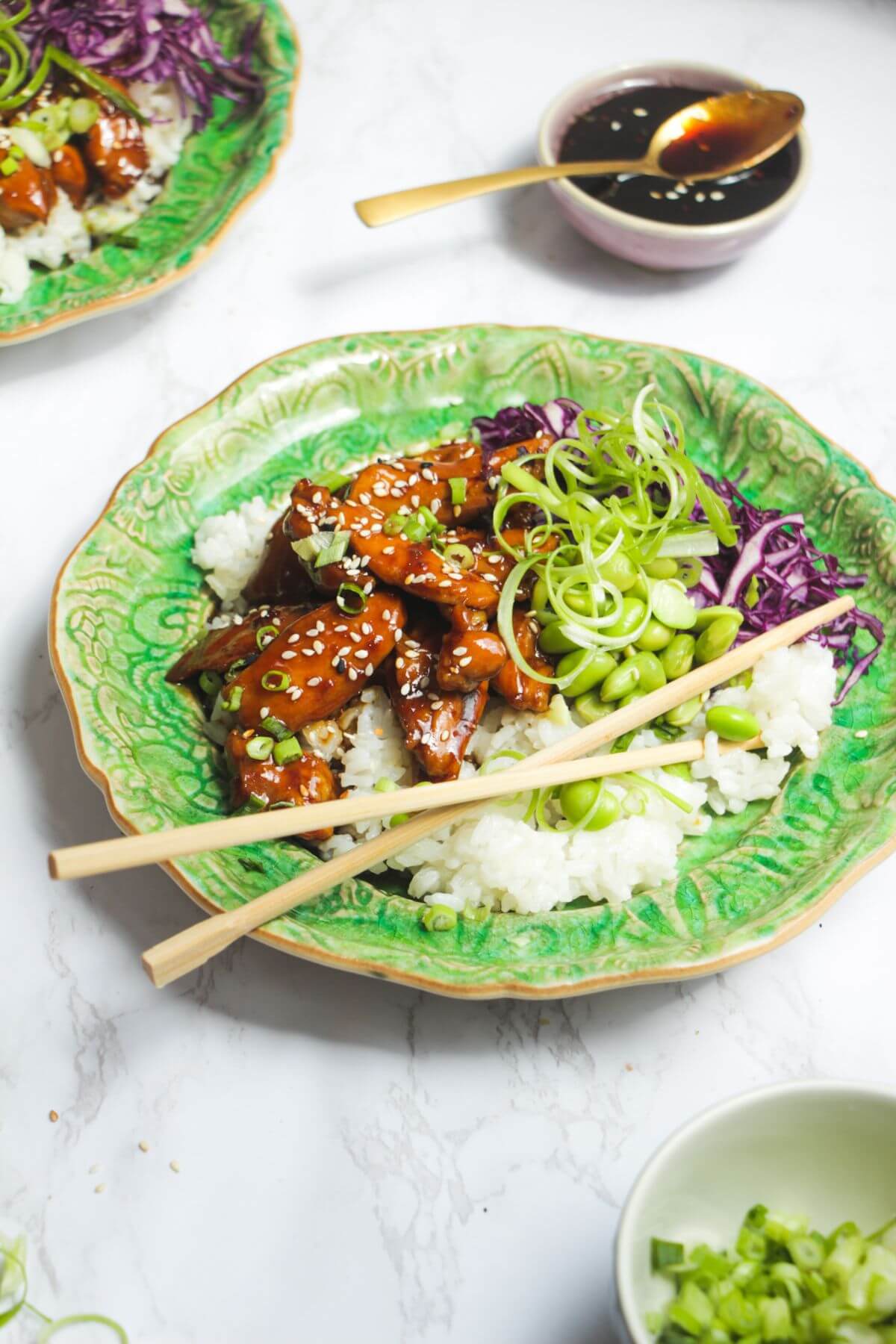 Glossy teriyaki chicken pieces on rice, with red cabbage and scallions on a green plate.