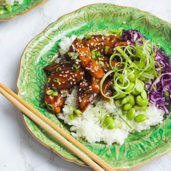 Glossy teriyaki chicken pieces on rice, with red cabbage and scallions on a green plate.