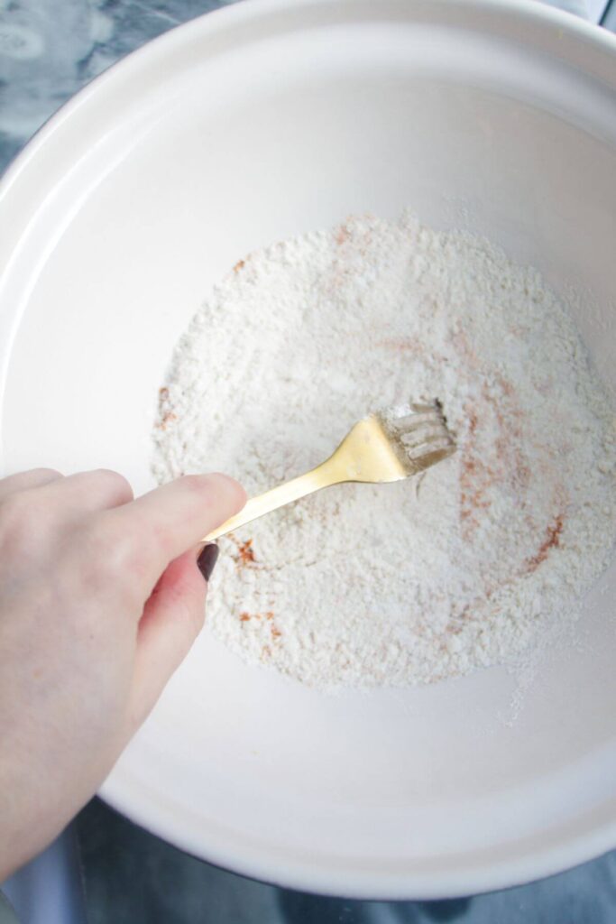 Hand holding a gold fork whisking flour and turmeric in a large white mixing bowl.
