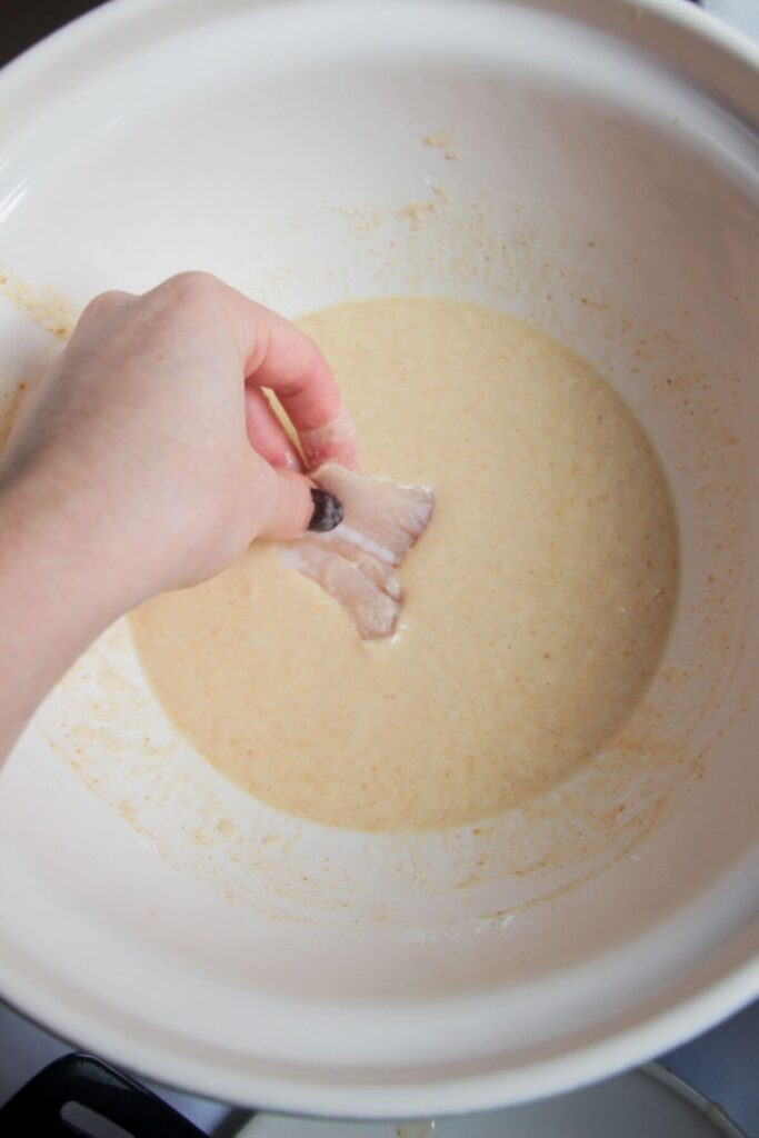 Hand dipping a piece of fish into wet batter in a large mixing bowl.