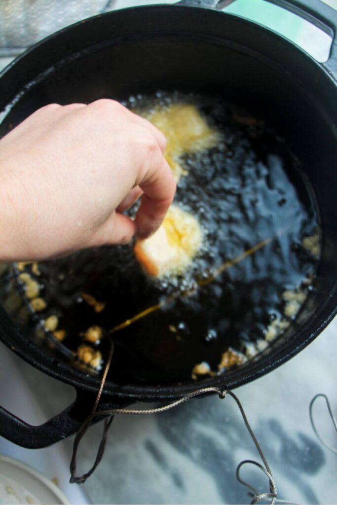 Hand lowering a piece of battered fish into hot oil in a large black pot.